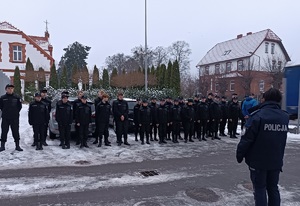 Plac przed komendą. Policjantka stoi przed uczniami ustawionymi w rzędzie.