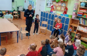 Sala lekcyjna. Policjantka stoi przed przedszkolakami, którzy siedzą na podłodze. Policjantka zwrócona jest przodem do zdjęcia. W tle widać półki z zabawkami.