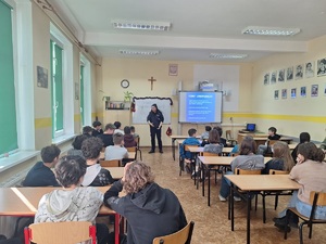 Policjantka prowadzi zajęcia w klasie z uczniami. Uczniowie siedzą w ławkach szkolnych, przed nimi stoi policjantka.