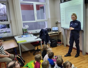 Policjantka stoi przed dziećmi siedzącymi na ławkach szkolnych. Za plecami wyświetla się prezentacja multimedialna dotycząca bezpiecznych ferii.