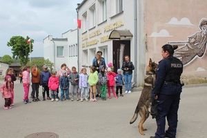 Policjantka razem z psem stoi przed dziećmi przed budynkiem biblioteki