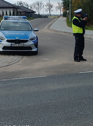 Policjant ruchu drogowego  stoi przy drodze . W ręku trzyma miernik prędkości