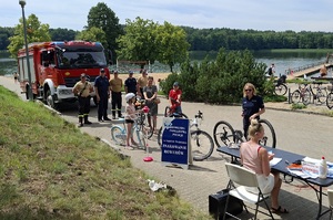 Policjantka znakuje rower. w oddali widać kolejkę chętnych do znakowania rowerów oraz pojazd straży pożarnej.