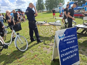 Policjant stoi przy rowerze ułożonym na ławce. Przygotowuje się do znakowania roweru. W tle widać uczestników pikniku.