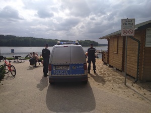 Policjanci obserwują kąpielisko. Stoją obok radiowozu. w oddali widać osoby korzystające z plaży.
