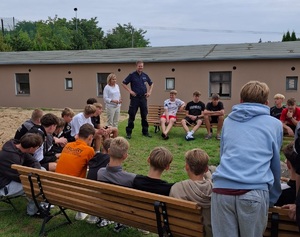 Policjant wspólnie z przedstawicielem Sanepidu stoi przed uczestnikami obozu sportowego. Wspólnie prowadzą spotkanie pod nazwa kręci mnie bezpieczeństwo w lecie.