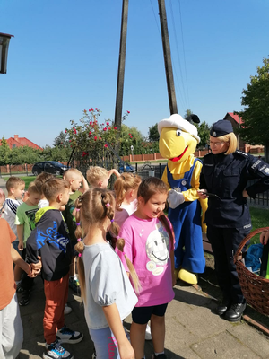 Maskotka mrówka, policjantka , uczniowie stoją przed budynkiem szkoły