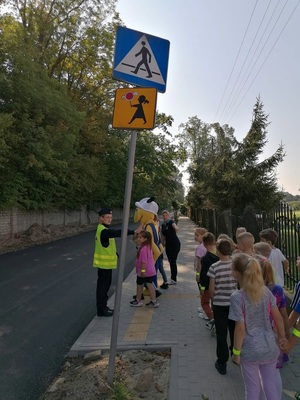Policjantka stoi przed przejściem razem z dziećmi i maskotka mrówką