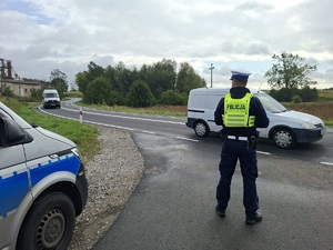 Policjant ruchu drogowego stoi przed jezdnia, która jada samochody. W ręku trzyma miernik prędkości.