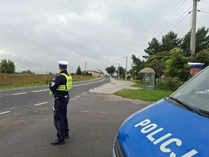 Policjant ruchu drogowego stoi przed jezdnia, którą jedzie samochód. W ręku trzyma miernik prędkości. Z prawej strony zdjęcia widać część zaparkowanego radiowozu.
