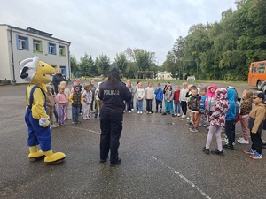 Policjantka rozmawia z dziećmi o zasadach bezpiecznej drogi do szkoły. Obok niej stoi maskotka Mrówki