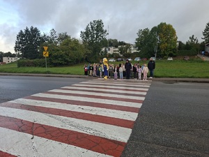 Policjantka rozmawia z dziećmi o zasadach bezpiecznej drogi do szkoły. Obok niej stoi maskotka Mrówki. Wszyscy stoją przed przejściem dla pieszych.
