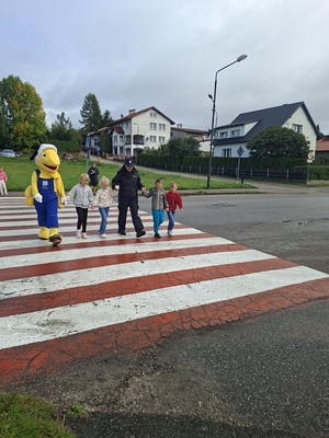 Policjantka wraz z dziećmi przechodzą przez przejście dla pieszych. Obok stoi maskotka Mrówki