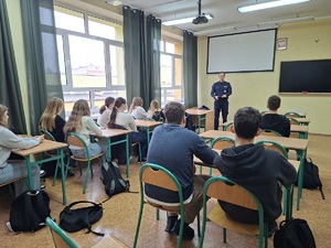 Dzień Promocji Zdrowia w Liceum ogólnokształcącym w Sępólnie Krajeńskim. Policjant stoi przed uczniami którzy siedzą w ławkach szkolnych. Uczniowie zwróceni są do zdjęcia plecami.