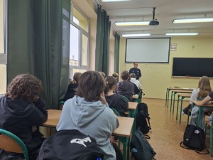 Dzień Promocji Zdrowia w Liceum ogólnokształcącym w Sępólnie Krajeńskim. Policjant stoi przed uczniami którzy siedzą w ławkach szkolnych. Uczniowie zwróceni są do zdjęcia plecami.