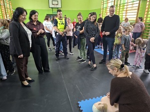 Sala gimnastyczna w szkole w Zamartem. Uczestnicy akcji bicia rekordu w udzielaniu pierwszej pomocy przypatrują się pani wykonującej uciski na fantomie. Pośród uczestników stoi policjant