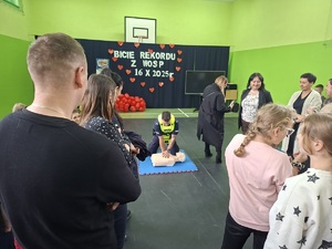 Sala gimnastyczna w szkole w Zamartem. W tle policjant który uczestniczy w akcji bicia rekordu w udzielaniu pierwszej pomocy pochyla się nad fantomem. Dookoła policjanta stoją inni uczestnicy akcji.