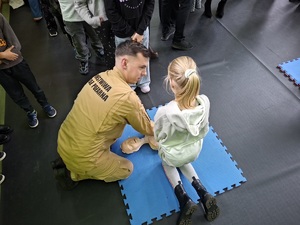 Sala gimnastyczna w szkole w Zamartem. Strażak udziela dziewczynce szkolenia prawidłowego wykonywania pierwszej pomocy na fantomie.