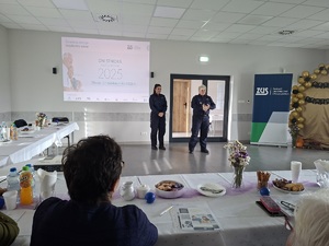 Prelekcja Policji z seniorami w Zbożu. Policjantki stoją przed seniorami na środku sali. Seniorzy siedzą przy stole. Policjantki są zwrócone twarzą do zdjęcia. W tle widać baner Policji i ZUS-u.
