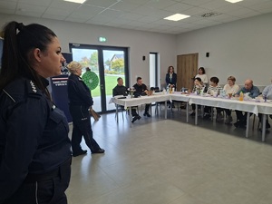 Prelekcja Policji z seniorami w Zbożu. Policjantki stoją przed seniorami na środku sali. Seniorzy siedzą przy stole.  W tle widać baner Policji i ZUS-u.