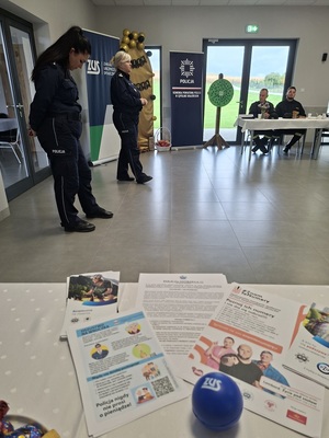Prelekcja Policji z seniorami w Zbożu. Policjantki stoją przed seniorami na środku sali. Seniorzy siedzą przy stole. Policjantki stoją bokiem do zdjęcia. W tle widać baner Policji i ZUS-u. Na pierwszym planie zdjęcia widać rozłożone na stole policyjne ulotki dotyczące bezpieczeństwa seniorów.