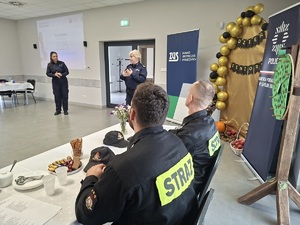 Prelekcja Policji z seniorami w Zbożu. Policjantki stoją przed seniorami na środku sali. Przy stole plecami do zdjęcia siedzą strażacy. Policjantki są zwrócone twarzą do zdjęcia. W tle widać baner Policji i ZUS-u.