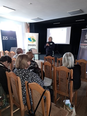 Prelekcja Policji z seniorami w Kamieniu Krajeńskim. Policjantki stoją przed seniorami na środku sali. Seniorzy siedzą na krzesłach. Zwróceni są plecami do zdjęciach  Policjantki są zwrócone twarzą do zdjęcia. W tle widać baner Policji i ZUS-u.
