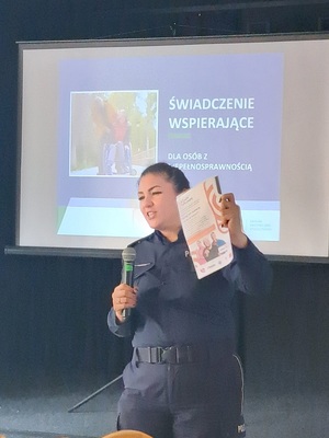 Prelekcja Policji z seniorami w Kamieniu Krajeńskim. Policjantka stoją przed seniorami na środku sali. Seniorzy siedzą na krzesłach. Zwróceni są plecami do zdjęcia. W tle widać baner Policji i ZUS-u.