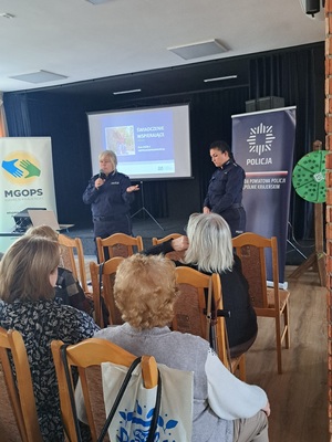 Prelekcja Policji z seniorami w Kamieniu Krajeńskim. Policjantki stoją przed seniorami na środku sali. Seniorzy siedzą na krzesłach. Zwróceni są plecami do zdjęcia.  Policjantki są zwrócone twarzą do zdjęcia. W tle widać baner Policji i ZUS-u.