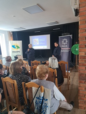 Prelekcja Policji z seniorami w Kamieniu Krajeńskim. Policjantki stoją przed seniorami na środku sali. Seniorzy siedzą na krzesłach. Zwróceni są plecami do zdjęcia.  Policjantki są zwrócone twarzą do zdjęcia. W tle widać baner Policji i ZUS-u.