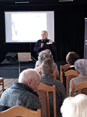 Prelekcja Policji z seniorami w Kamieniu Krajeńskim. Policjantka stoi przed seniorami na środku sali. Seniorzy siedzą na krzesłach. Zwróceni są plecami do zdjęcia.  Policjanta jest zwrócona twarzą do zdjęcia. W tle widać baner Policji i ZUS-u.
