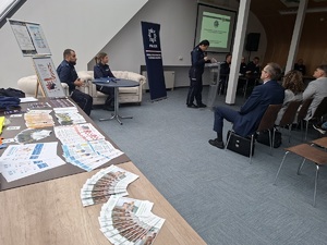 Debata społeczna pn. „Bezpieczeństwo seniorów we współczesnym świecie manipulacji oszustów”. Po prawej stronie widać uczestników debaty siedzących w sali konferencyjnej na krzesłach ustawionych w rzędach. Zwróceni są plecami  do zdjęcia . Przed nimi przy stole siedzą policjanci. W tle wyświetlana jest prezentacja multimedialna. Z lewej strony zdjęcia na stole widać rozłożone ulotki.