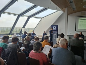 Debata społeczna pn. „Bezpieczeństwo seniorów we współczesnym świecie manipulacji oszustów”. Uczestnicy debaty siedzą w sali konferencyjnej na krzesłach ustawionych w rzędach. Zwróceni są do zdjęcia plecami. Przed nimi przy stole siedzą policjanci. W tle wyświetlana jest prezentacja multimedialna.