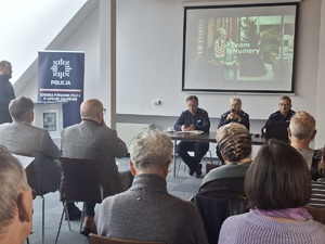 Debata społeczna pn. „Bezpieczeństwo seniorów we współczesnym świecie manipulacji oszustów”. Uczestnicy debaty siedzą w sali konferencyjnej na krzesłach ustawionych w rzędach. Zwróceni są do zdjęcia plecami. Przed nimi przy stole siedzą policjanci. W tle wyświetlany jest film .