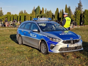 Policjant stoi przy radiowozie . W oddali widać cmentarz