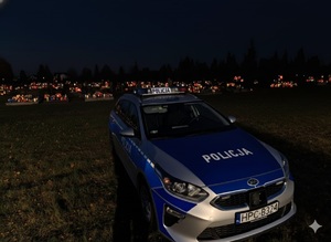 Jest noc. Policyjny radiowóz stoi zaparkowany przed cmentarzem. w oddali widać palące się znicze.