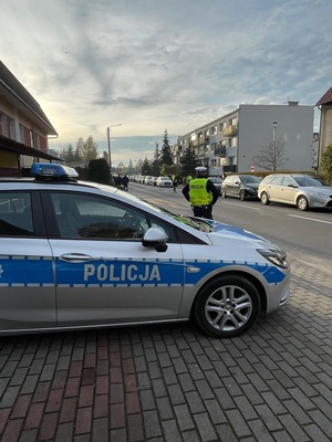 Policjant stoi przed zaparkowanym radiowozem. w oddali widać zaparkowane pojazdy oraz budynki wielorodzinne