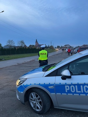 Policjant stoi przed zaparkowanym radiowozem. w oddali widać zaparkowane pojazdy oraz przemieszczających się pieszych