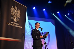 Policjant stoi przed mikrofonem. W tle widać wyświetlany napis 100-lat Kobiet w Policji. Po lewej stronie widać baner Wojewody Kujawsko-Pomorskiego.
