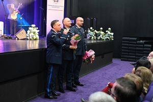 Policjanci stoją pod sceną. W rękach trzymają kwiaty. Zwróceni są przodem do widowni.