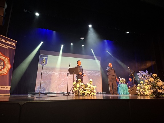 Sala kinowa w Centrum Kultury i Sztuki w Sępólnie Krajeńskim. Na scenie śpiewa dwóch policjantów, a za ich plecami na sofie siedzą dwie policjantki oraz pracownik cywilny. W tle wyświetlany jest film promujący poszczególne gminy powiatu sępoleńskiego. Po bokach sceny  ustawione są banery promujące Komendę Powiatowa Policji w Sępólnie Krajeńskim oraz Niezależny Samorządny Związek Zawodowy Policjantów w Sępólnie Krajeńskim