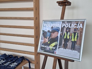 Zdjęcie plakatu promującego zawód policjanta z napisem Dołącz do Nas umieszczony w ramce  ustawionej na stojaku