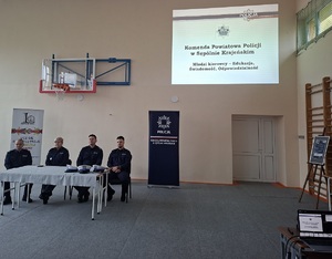 Policjanci podczas debaty siedzący przy stole. Za ich plecami na ścianie wyświetlana jest prezentacja multimedialna z napisem „Młodzi kierowcy – edukacja, świadomość, odpowiedzialność”