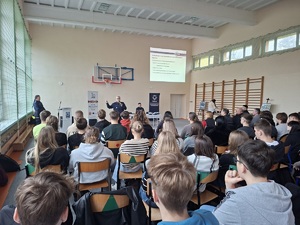 zdjęcie uczestników debaty siedzących na krzesłach. Zwróceni są plecami do zdjęcia . W oddali widać policjantów siedzących przed uczestnikami debaty. W tle wyświetlana jest na ścianie prezentacja multimedialna.