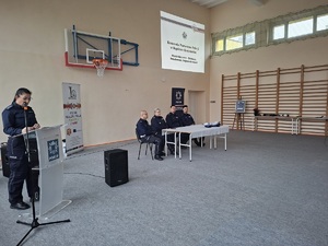 Zdjęcie policjantów uczestniczących w debacie. Przy mównicy stoi policjantka, a pozostali policjanci siedzą po jej lewej stronie przy stole. W tle na ścianie wyświetlana jest prezentacja multimedialna  z napisem „Młodzi kierowcy – edukacja, świadomość, odpowiedzialność”