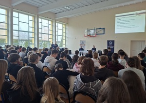 zdjęcie uczestników debaty siedzących na krzesłach. Zwróceni są plecami do zdjęcia . W oddali widać policjantów siedzących przodem do uczestnikami debaty. W tle wyświetlana jest na ścianie prezentacja multimedialna.