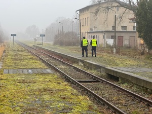 Zdjęcie policjanta oraz pracownika Służby Ochrony Kolei w trakcie patrolu wzdłuż torów kolejowych. W oddali widać budynki oraz drzewa.