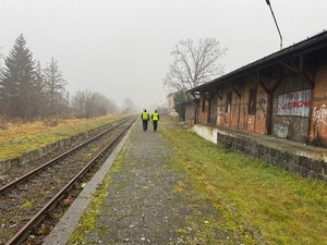 Zdjęcie policjanta oraz pracownika Służby Ochrony Kolei w trakcie patrolu wzdłuż torów kolejowych. W oddali widać budynki oraz drzewa.