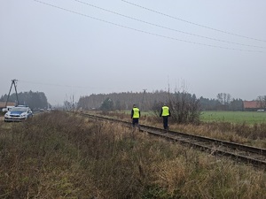 Zdjęcie policjantów w trakcie patrolu wzdłuż torów kolejowych. W oddali widać radiowóz policyjny, zabudowania oraz drzewa.