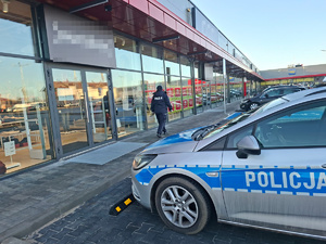 Policjantka idzie wzdłuż pasażu handlowego. Na parkingu widać zaparkowany radiowóz.